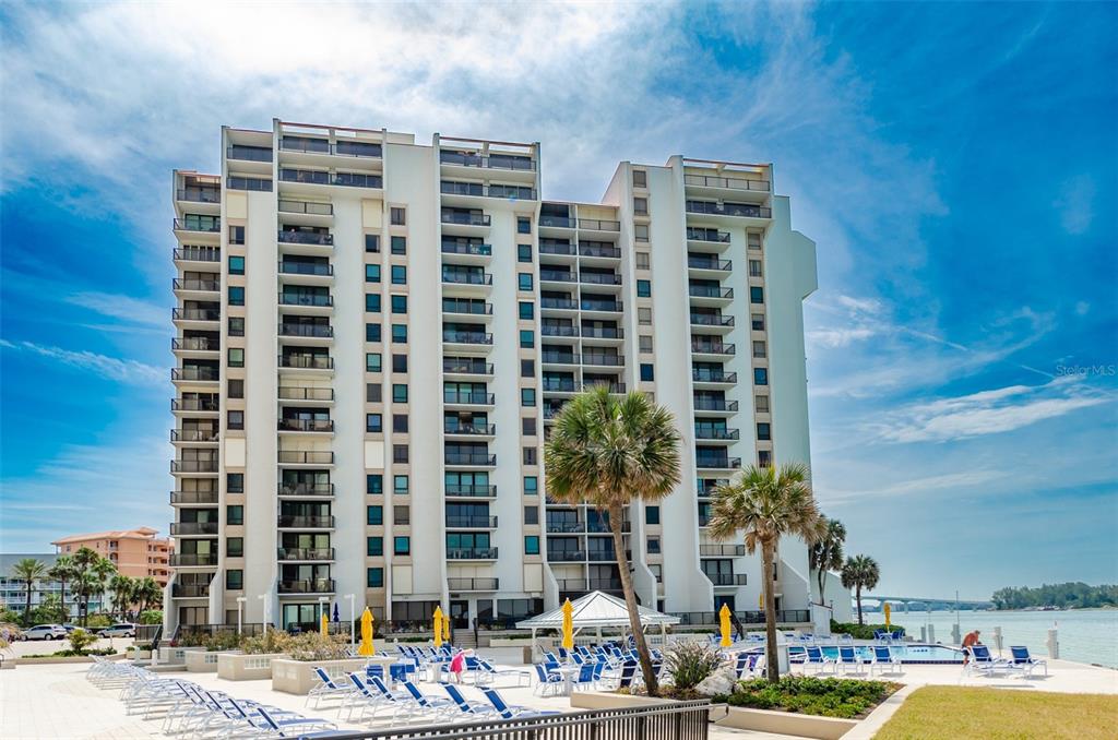 A tall white condominium building stands beside a swimming pool with lounge chairs and palm trees — 450 S GULFVIEW BOULEVARD UNIT 405, CLEARWATER, FL