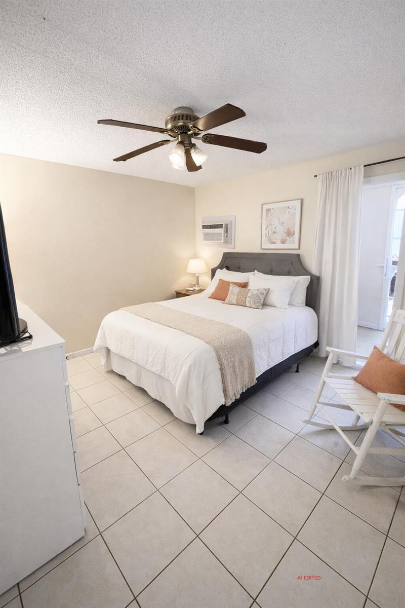 A bedroom features a bed, ceiling fan, white tile floors, and a white rocking chair — 7 Lyndhurst A, Deerfield Beach FL 33442, DEERFIELD BEACH, FL