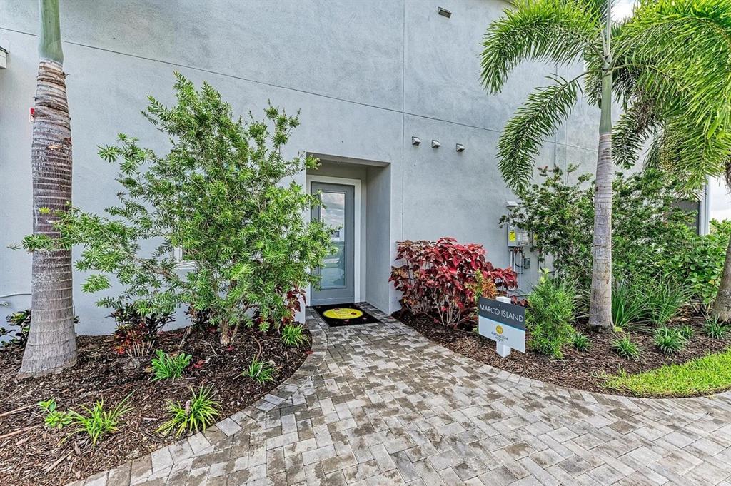 A paved walkway leads to a gray front door flanked by landscaping and palm trees — 10016 REMBRANDT WAY UNIT 101, BRADENTON, FL