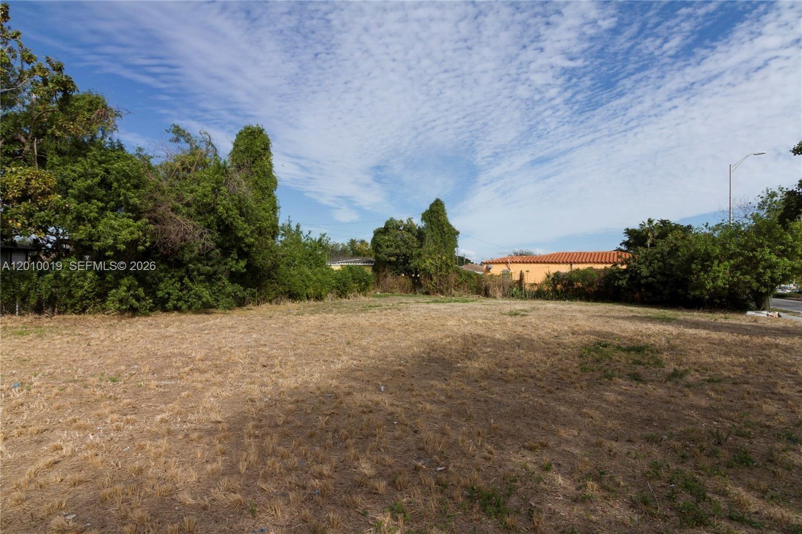 A vacant, dry grassy lot is bordered by dense trees and a neighboring orange house — 1765 NW 62nd St, Miami FL 33147, MIAMI, FL