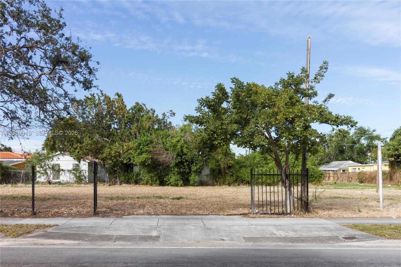 A vacant, fenced residential lot features a sidewalk in the foreground and mature trees — 1765 NW 62nd St, Miami FL 33147, MIAMI, FL