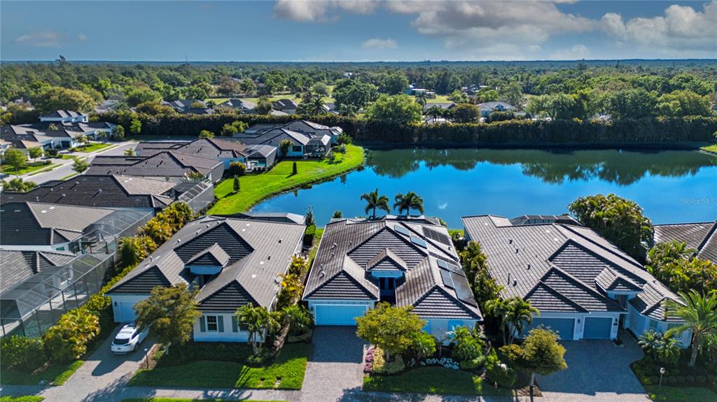 An aerial view shows three suburban houses with dark tiled roofs bordering a large pond — 7732 SANDHILL LAKE DRIVE, SARASOTA, FL