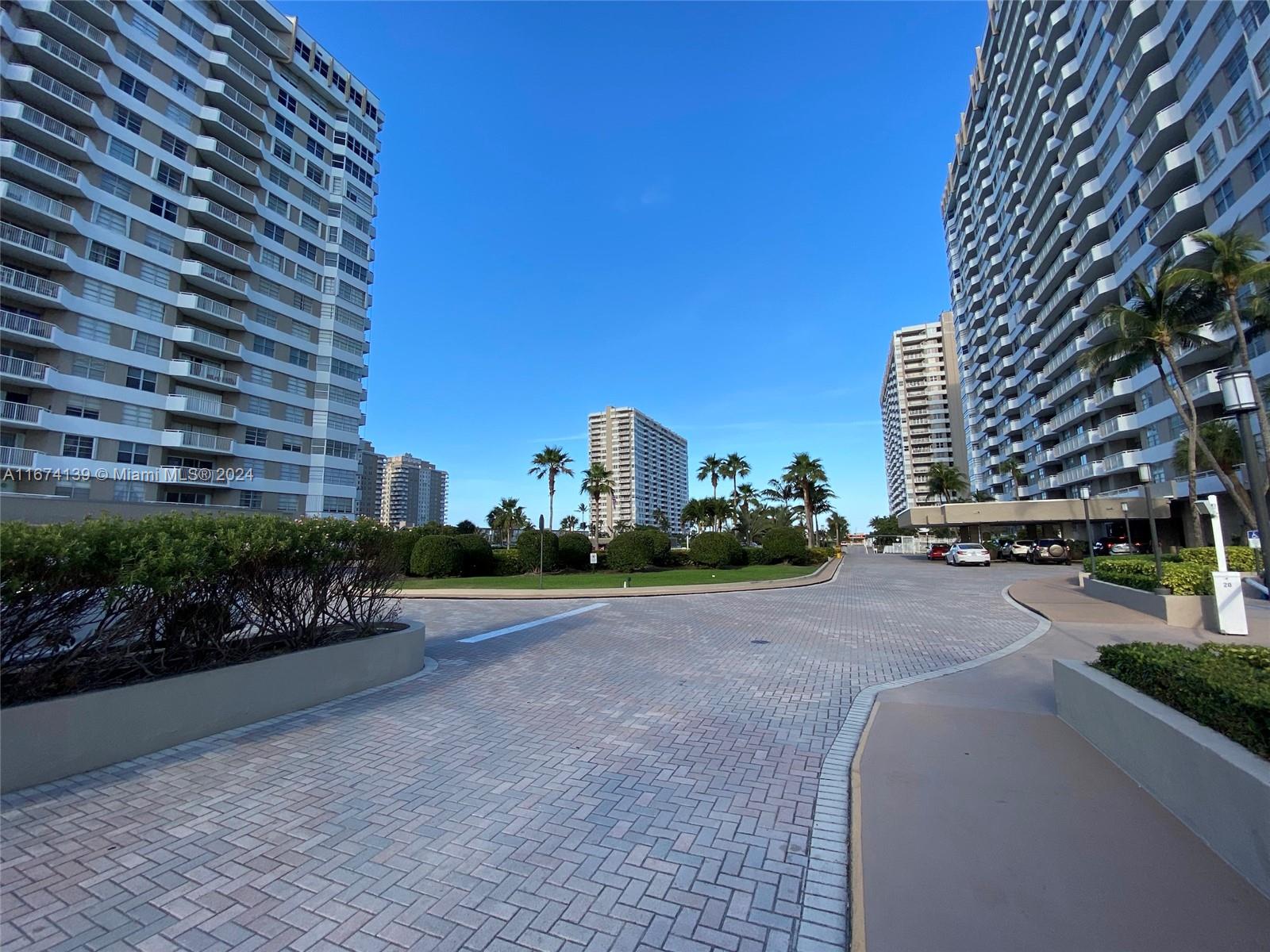 A paved driveway leads toward high-rise apartment buildings surrounded by palm trees and landscaping — 1965 S Ocean Dr # 4D, Hallandale Beach FL 33009, HALLANDALE BEACH, FL