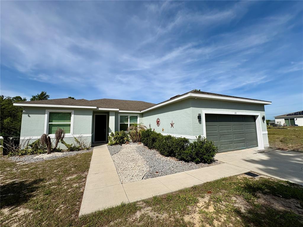A light green single-story house with a dark garage door and a concrete walkway — 65 ZINNIA LANE E, KISSIMMEE, FL