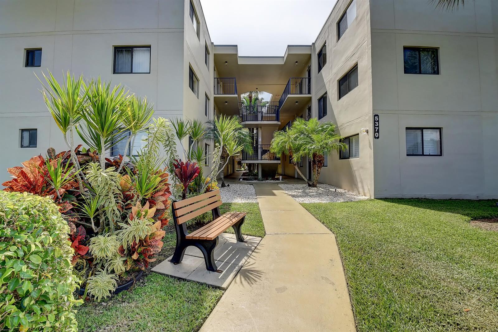 A concrete walkway leads to the entrance of a multi-story apartment building with landscaping — 5370 Las Verdes Circle # 324, Delray Beach FL 33484, DELRAY BEACH, FL