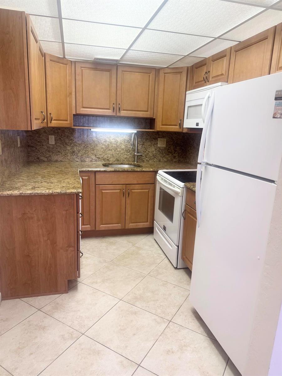 A kitchen features wood cabinets, granite countertops, tile flooring, and white appliances — 2381 Sunset Avenue # 117, FL 33461, FL