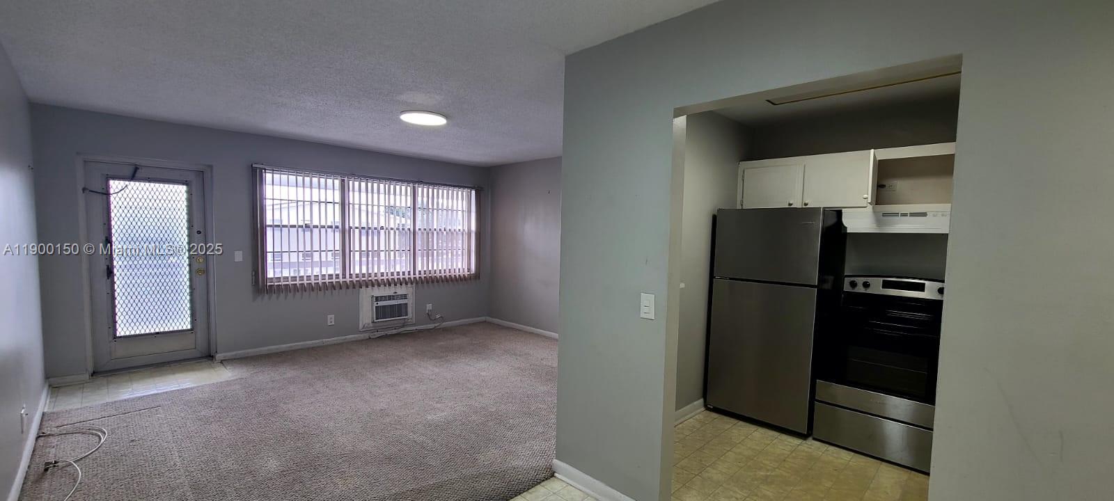 A carpeted living room with a window and a small adjacent kitchen with stainless appliances — 92 Berkshire D # 92, West Palm Beach FL 33417, WEST PALM BEACH, FL