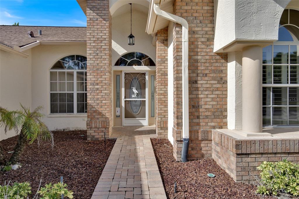 A brick-accented house exterior features a paved walkway leading to a glass-paned front door — 18601 QUEEN BROOKS COURT, HUDSON, FL