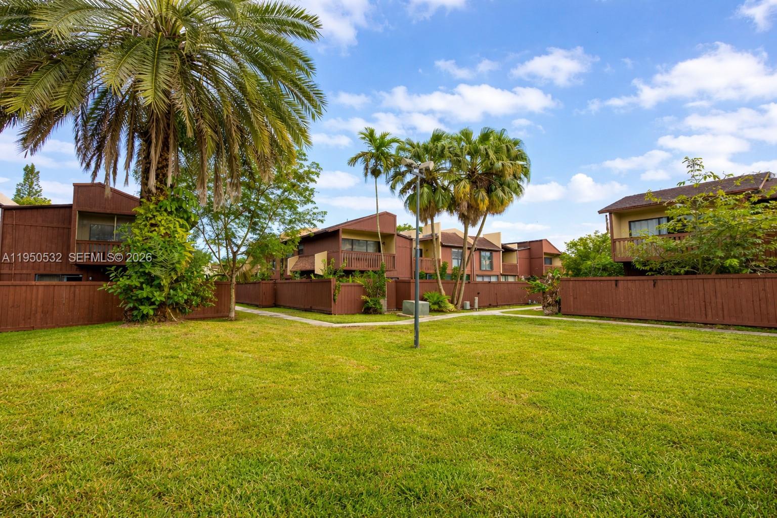 A grassy courtyard sits between brown multi-story residential buildings with wooden fences and palm trees — 6534 SW 114th Pl # 75, Miami FL 33173, MIAMI, FL