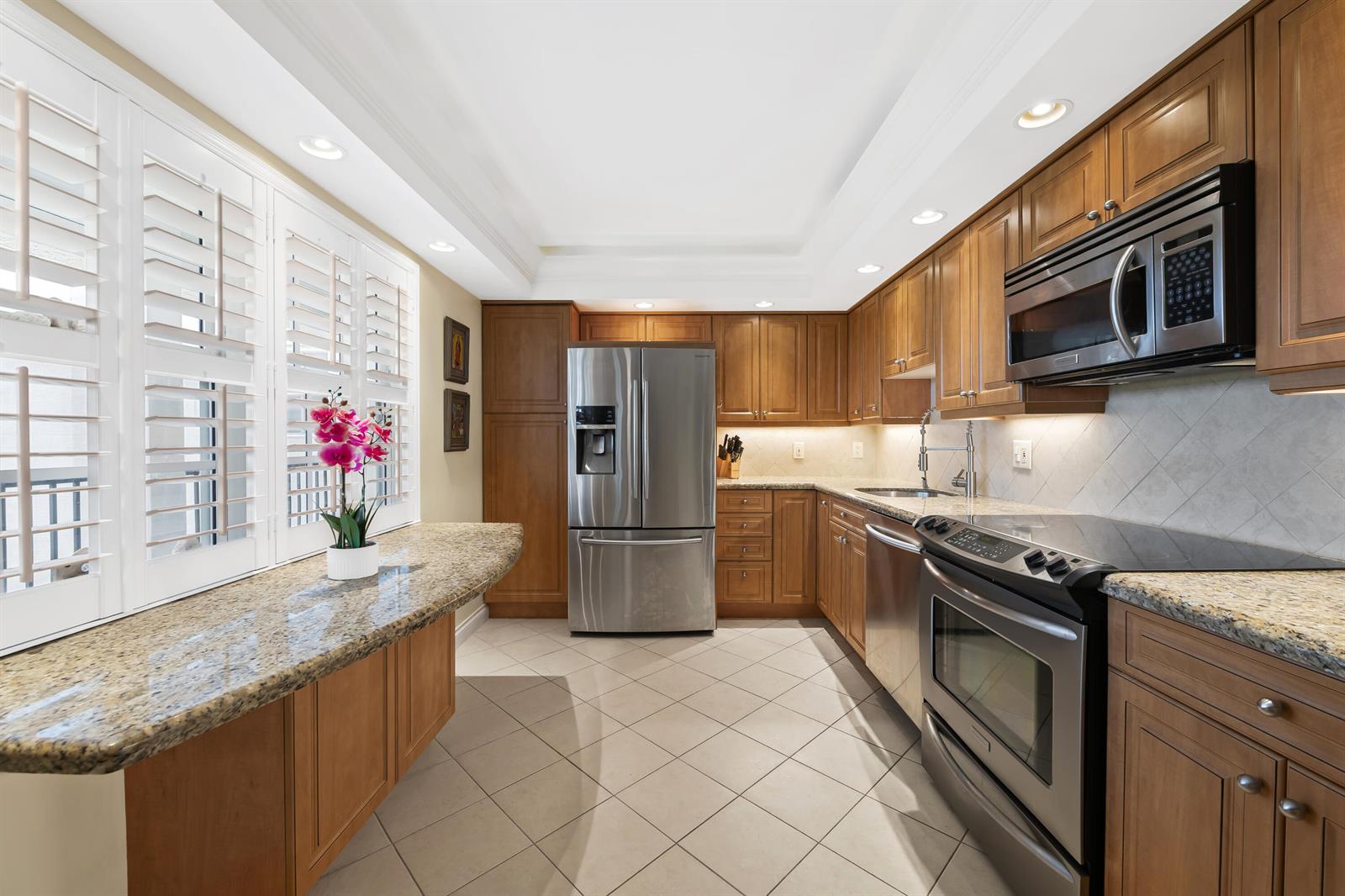 A kitchen features wood cabinets, granite countertops, stainless steel appliances, and a window with shutters — 2900 N Course Drive # 909, Pompano Beach FL 33069, POMPANO BEACH, FL
