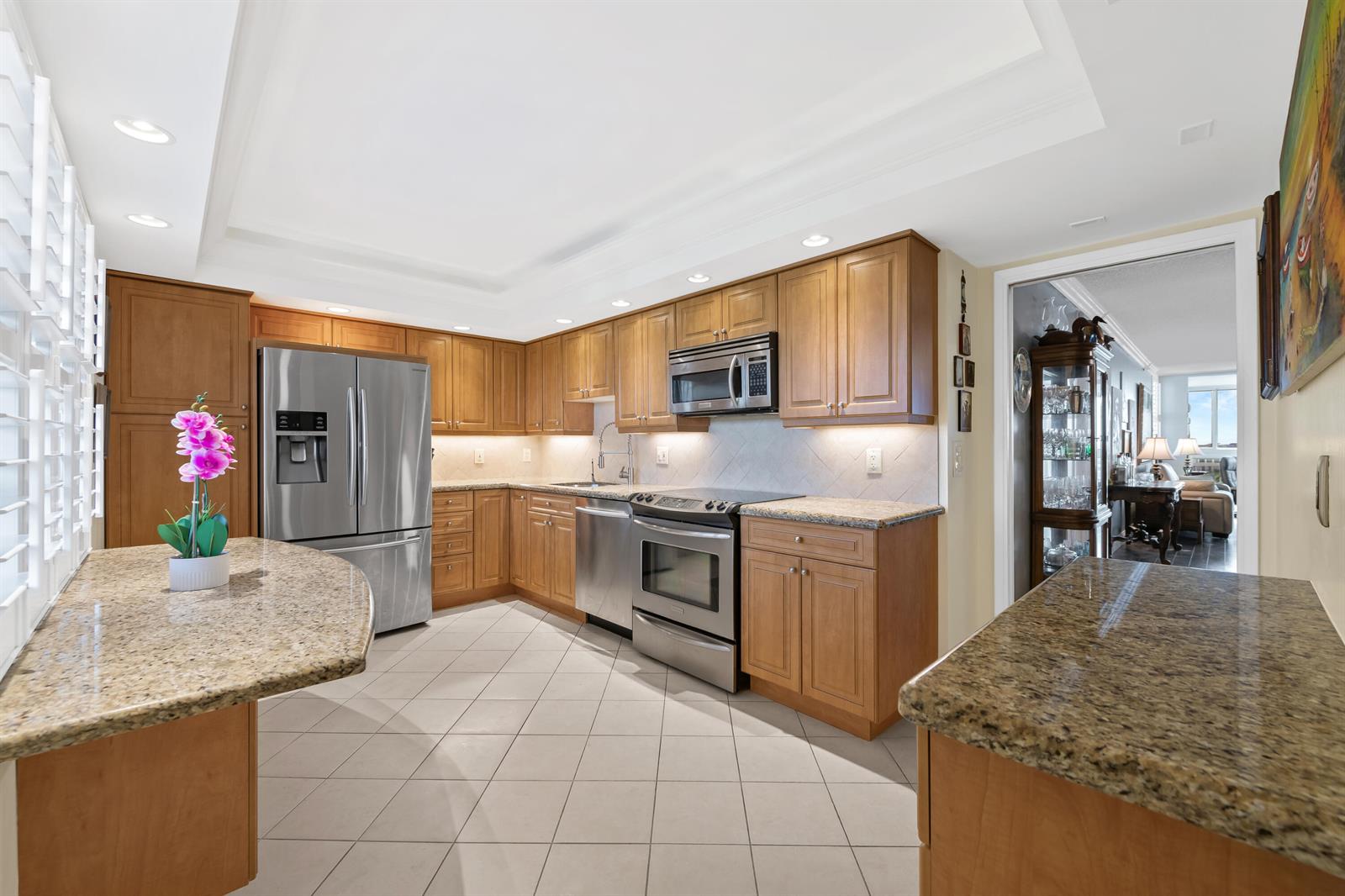 A kitchen features light wood cabinets, granite countertops, stainless steel appliances, and tile flooring — 2900 N Course Drive # 909, Pompano Beach FL 33069, POMPANO BEACH, FL