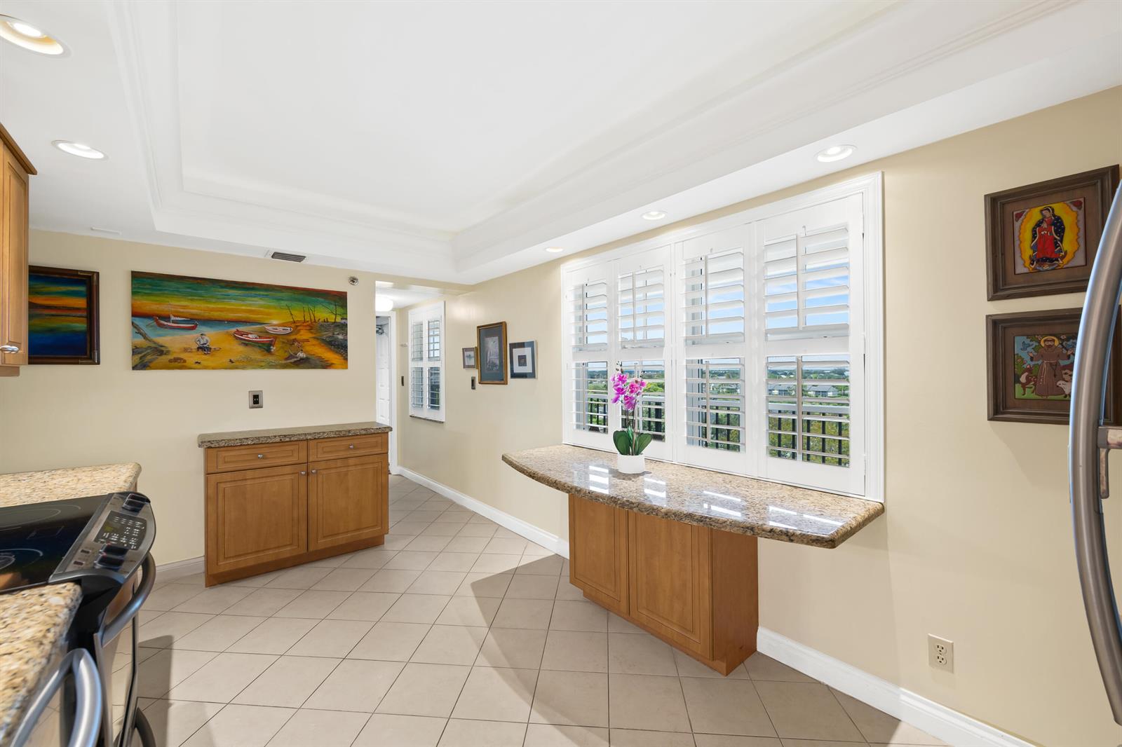 A kitchen features light wood cabinets, granite countertops, tile flooring, and plantation shutters on windows — 2900 N Course Drive # 909, Pompano Beach FL 33069, POMPANO BEACH, FL
