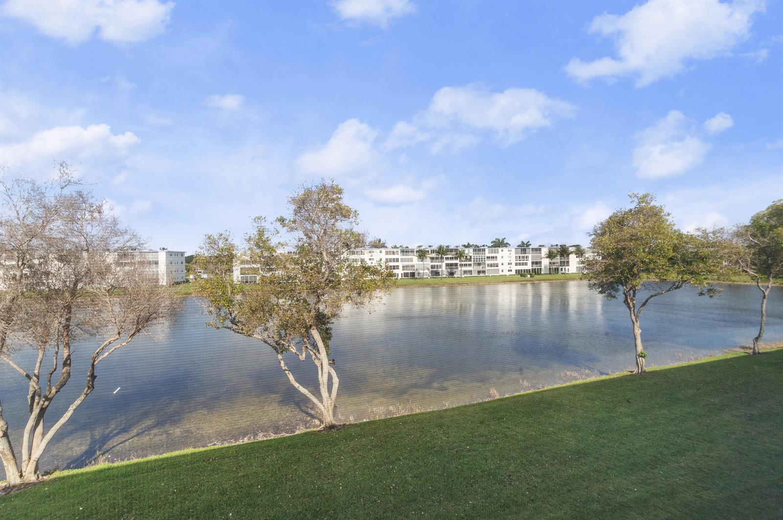 A grassy lawn overlooks a lake with apartment buildings in the background under a blue sky — 3013 Wolverton A, Boca Raton FL 33434, BOCA RATON, FL