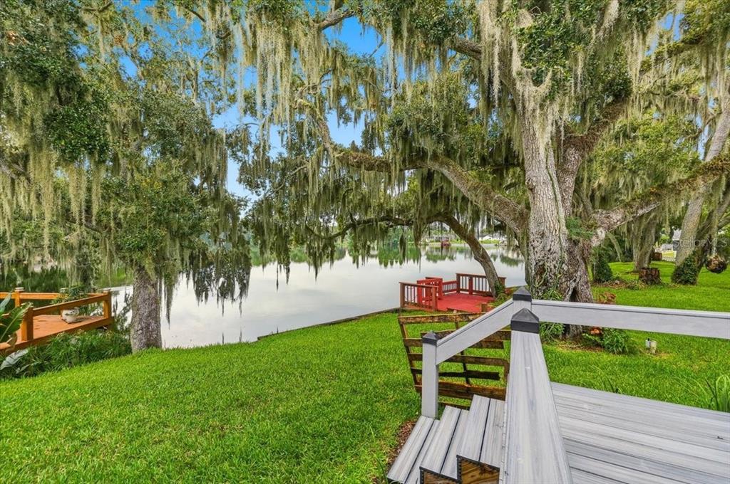 A grassy backyard slopes toward a lake, featuring large oak trees and two wooden decks — 6760 LAKE CLARK DRIVE, LAKELAND, FL