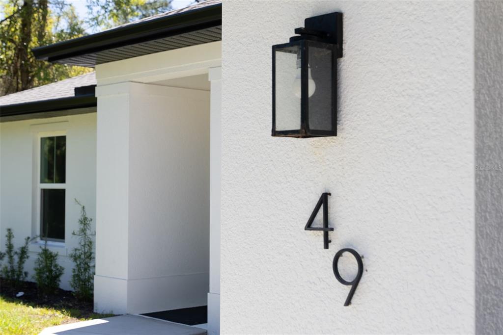 A close-up view of a white stucco exterior wall featuring a black light fixture and house numbers — 49 TROUTDALE STREET, PORT CHARLOTTE, FL