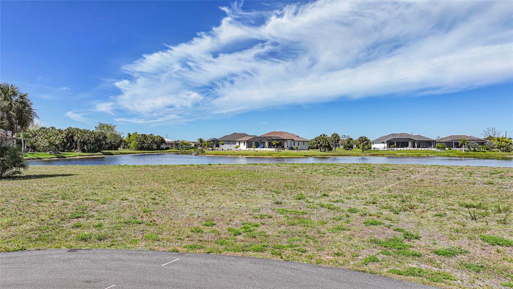 A vacant grassy lot overlooks a pond with houses in the background under a blue sky — 16395 NOGALES COURT, PUNTA GORDA, FL
