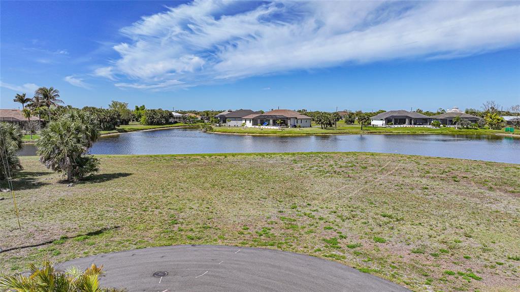 A vacant grassy lot overlooks a pond with houses in the background under a blue sky — 16395 NOGALES COURT, PUNTA GORDA, FL