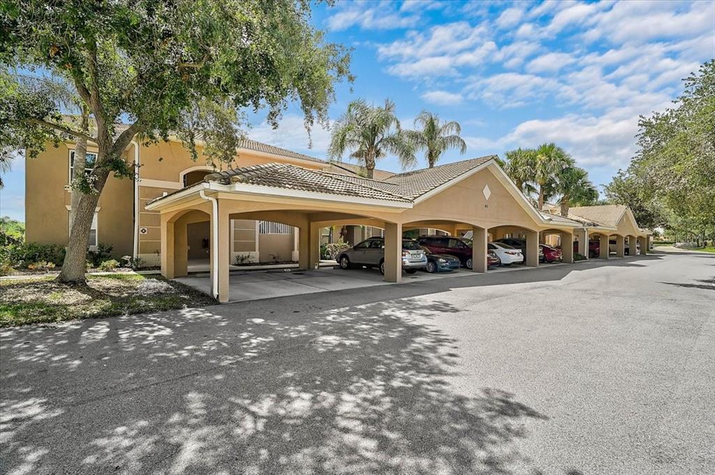 A tan apartment building features a long covered parking structure with several parked cars — 915 FAIRWAYCOVE LANE UNIT 201, BRADENTON, FL