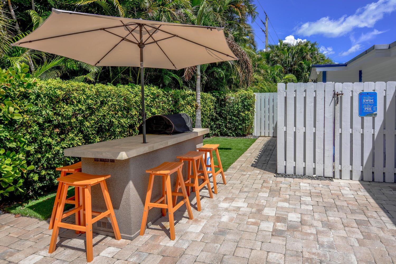 An outdoor kitchen with a stone bar, four wooden stools, and a patio umbrella — 3396 Pebble Place, Jupiter FL 33469, JUPITER, FL