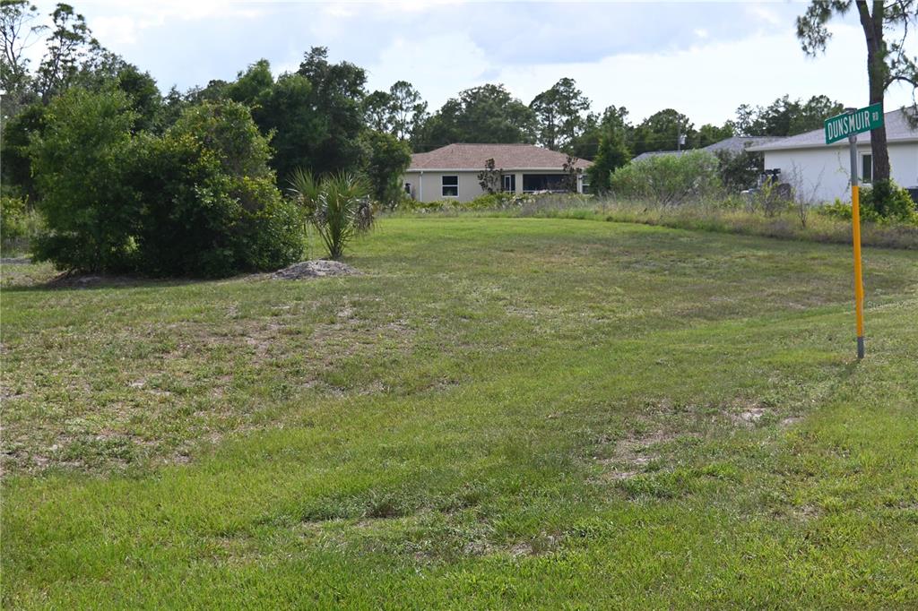 A vacant, grassy residential lot features a street sign labeled Dunsmuir Road — CHARMER LANE, NORTH PORT, FL