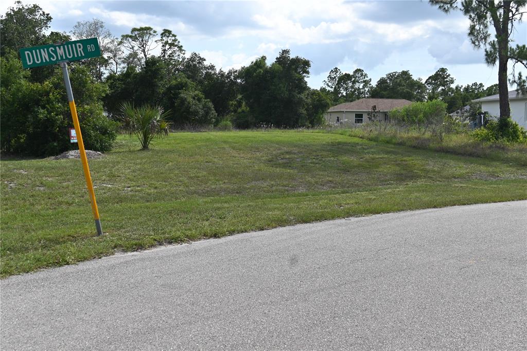A vacant, grassy residential lot sits at a street corner marked by a road sign — CHARMER LANE, NORTH PORT, FL