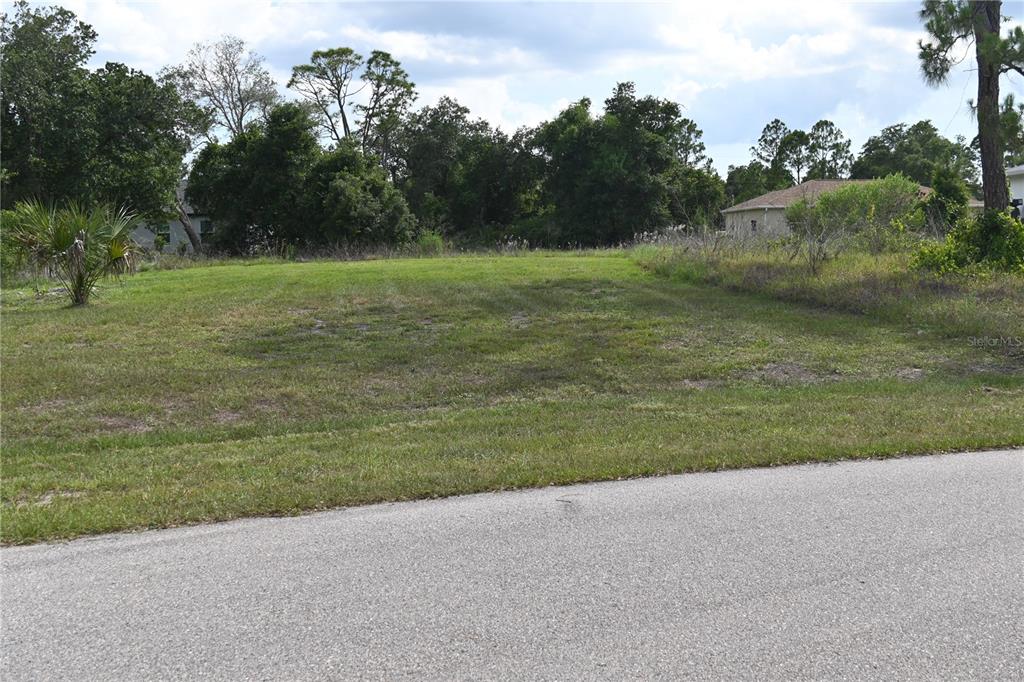 A grassy, vacant residential lot sits adjacent to a paved road with trees in the background — CHARMER LANE, NORTH PORT, FL