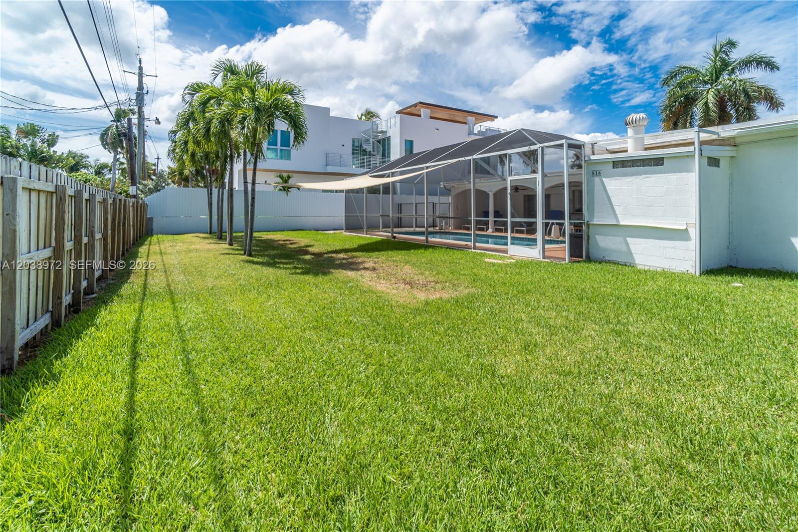 A grassy backyard features a screened-in swimming pool, palm trees, and a wooden fence — 816 Polk St # 1, Hollywood FL 33019, HOLLYWOOD, FL