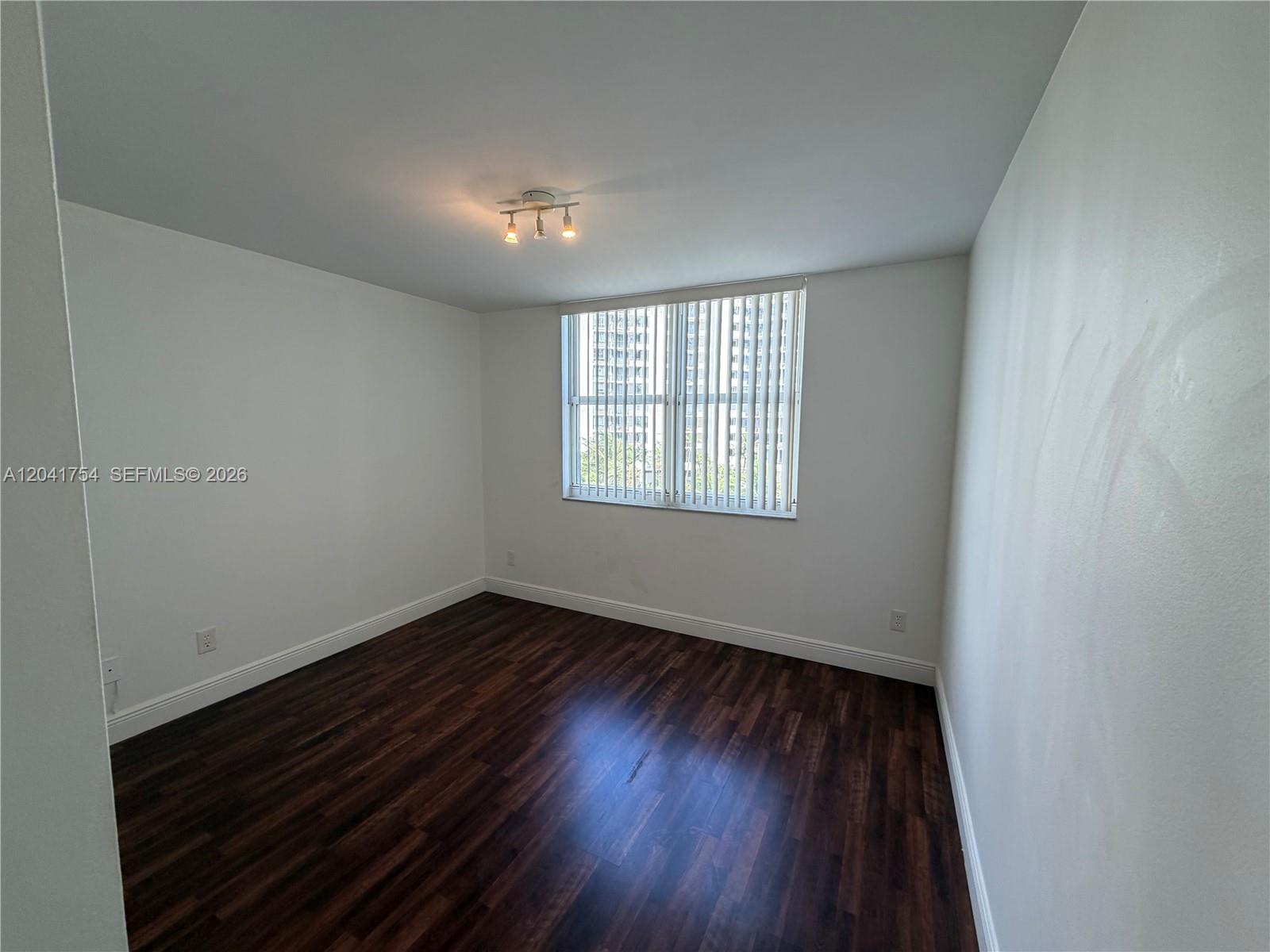 An empty room features dark wood-look flooring, white walls, and a window with vertical blinds — 770 Claughton Island Dr # 707, Miami FL 33131, MIAMI, FL