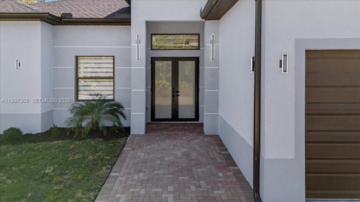 The exterior of a modern home features a brick walkway leading to double glass doors — 1504 E 6th, Lehigh Acres FL 33972, LEHIGH ACRES, FL