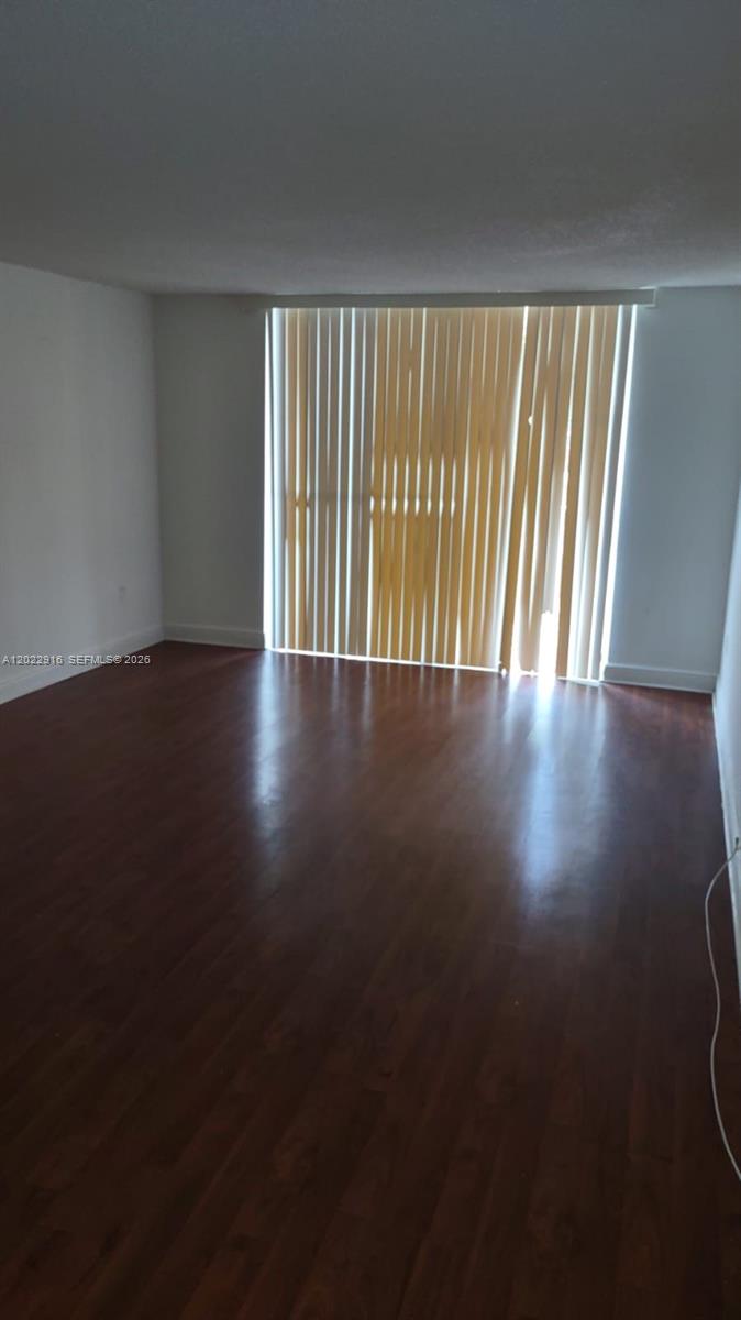 An empty room features dark wood-look flooring, white walls, and vertical blinds covering a window — 496 NW 165th St Rd # D109, Miami FL 33169, MIAMI, FL