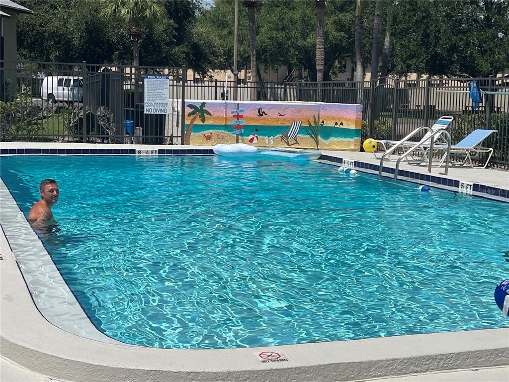 A man swims in a rectangular outdoor pool with a painted mural on the wall — 4215 E BAY DRIVE UNIT 1702H, CLEARWATER, FL