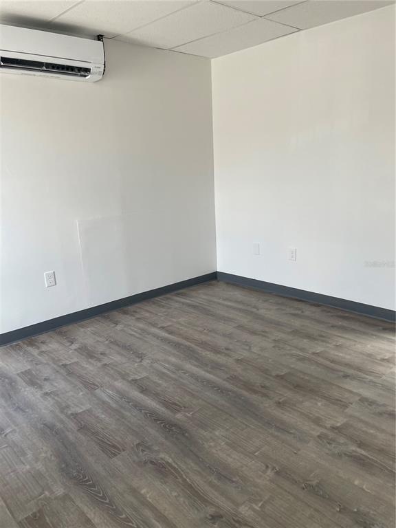 An empty room features white walls, dark gray baseboards, wood-look flooring, and a wall-mounted AC unit — 5525 COMMERCE DRIVE UNIT 3/4, ORLANDO, FL