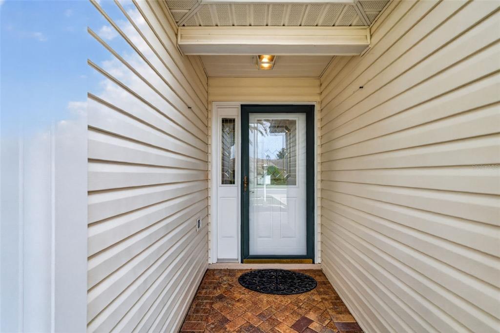 A front entryway with light-colored vinyl siding, a brick floor, and a glass-paneled front door — 1557 OAK FOREST DRIVE, THE VILLAGES, FL