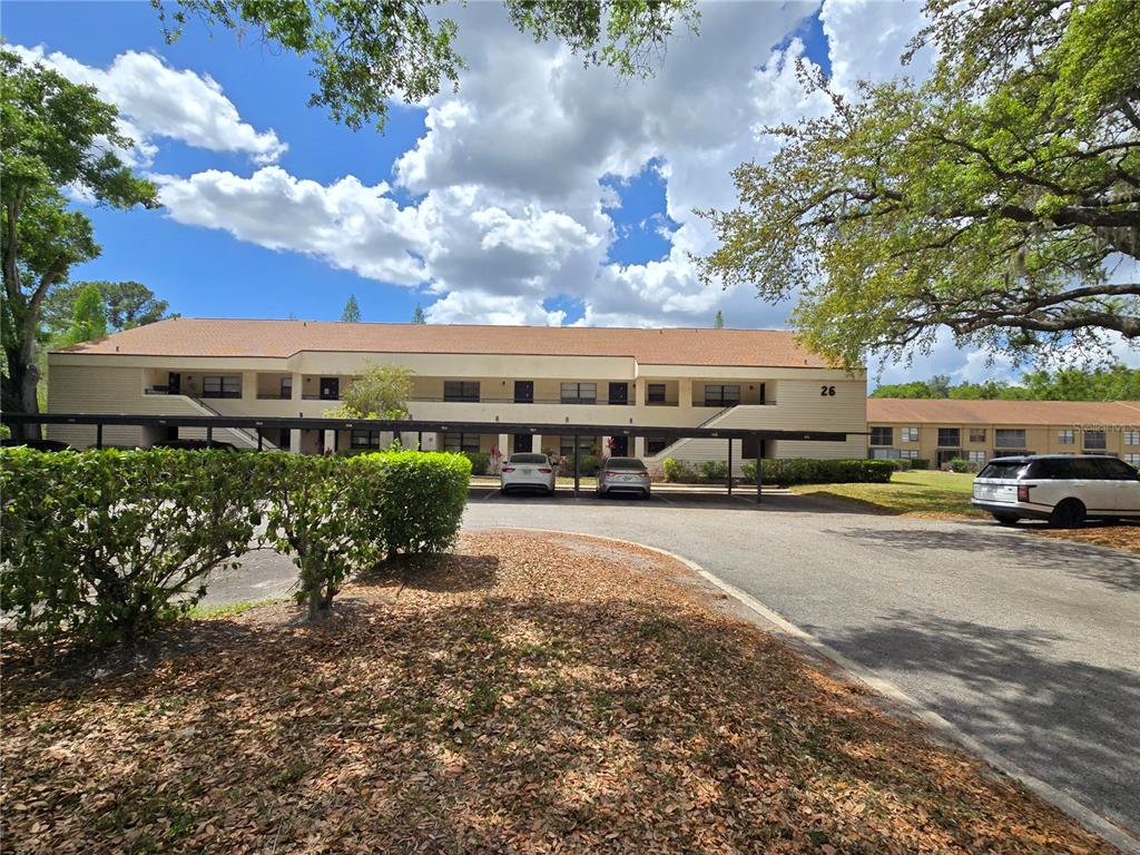 A two-story apartment building with covered parking spaces and mature trees in the foreground — 7838 NIAGARA AVENUE UNIT 305, TAMPA, FL