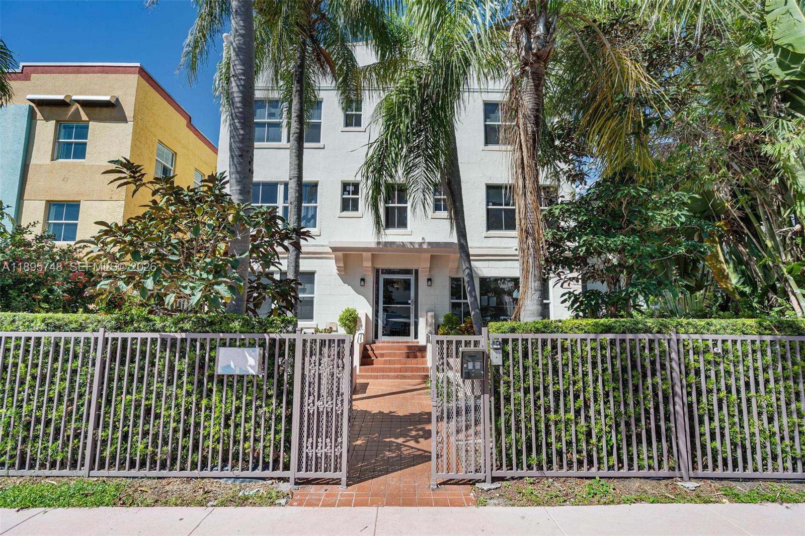 A three-story white apartment building stands behind a metal fence and palm trees — 1320 Drexel Ave # 200, Miami Beach FL 33139, MIAMI BEACH, FL