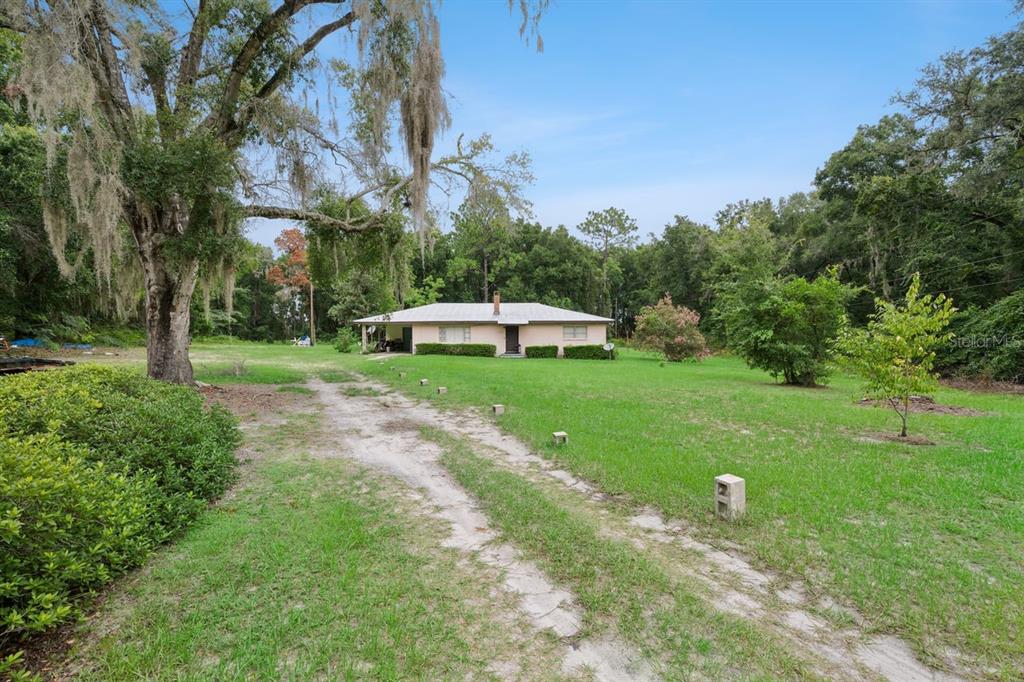 A single-story house sits on a large grassy lot with a dirt driveway and trees — 6184 NE 54TH BOULEVARD, WILDWOOD, FL