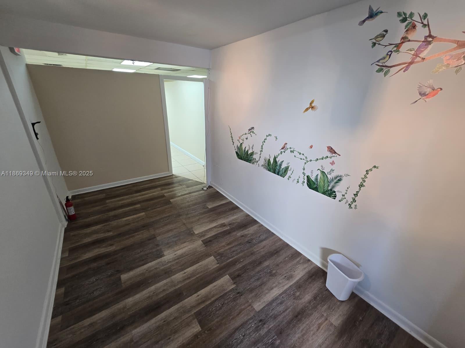 An empty room features wood-look flooring, a white wall with bird and plant decals, and a trashcan — 2501 N State Road 7 # 208, Margate FL 33063, MARGATE, FL