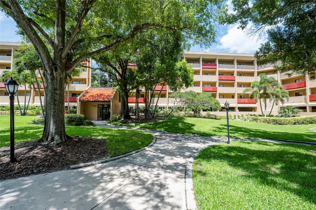 A concrete path leads to the entrance of a multi-story apartment building with red accents — 3062 EASTLAND BOULEVARD UNIT 406, CLEARWATER, FL