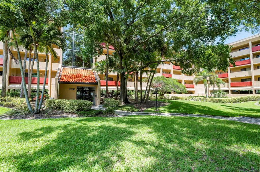 A multi-story apartment building with a terracotta-tiled entrance and surrounding palm trees and lawn — 3062 EASTLAND BOULEVARD UNIT 406, CLEARWATER, FL