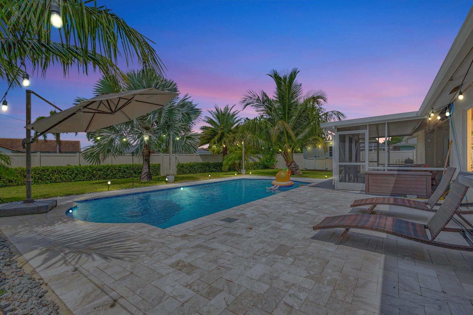 A backyard features a swimming pool, stone patio, lounge chairs, and palm trees at sunset — 1104 Jackpine Street, Wellington FL 33414, WELLINGTON, FL