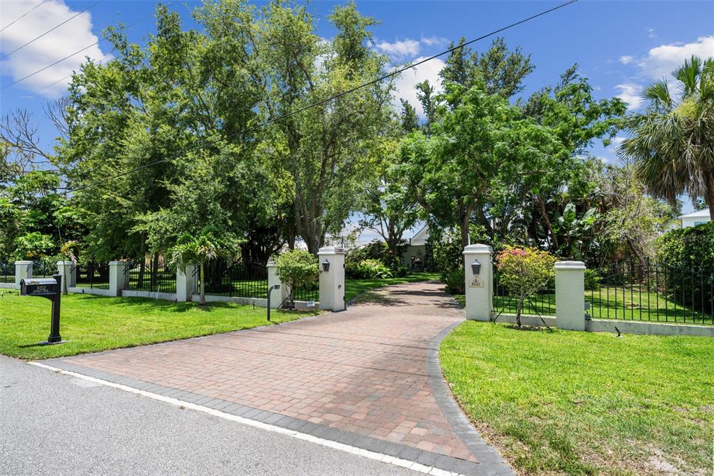 A brick driveway leads through a gated entrance to a house surrounded by mature trees — 8152 LONGBAY BOULEVARD, SARASOTA, FL