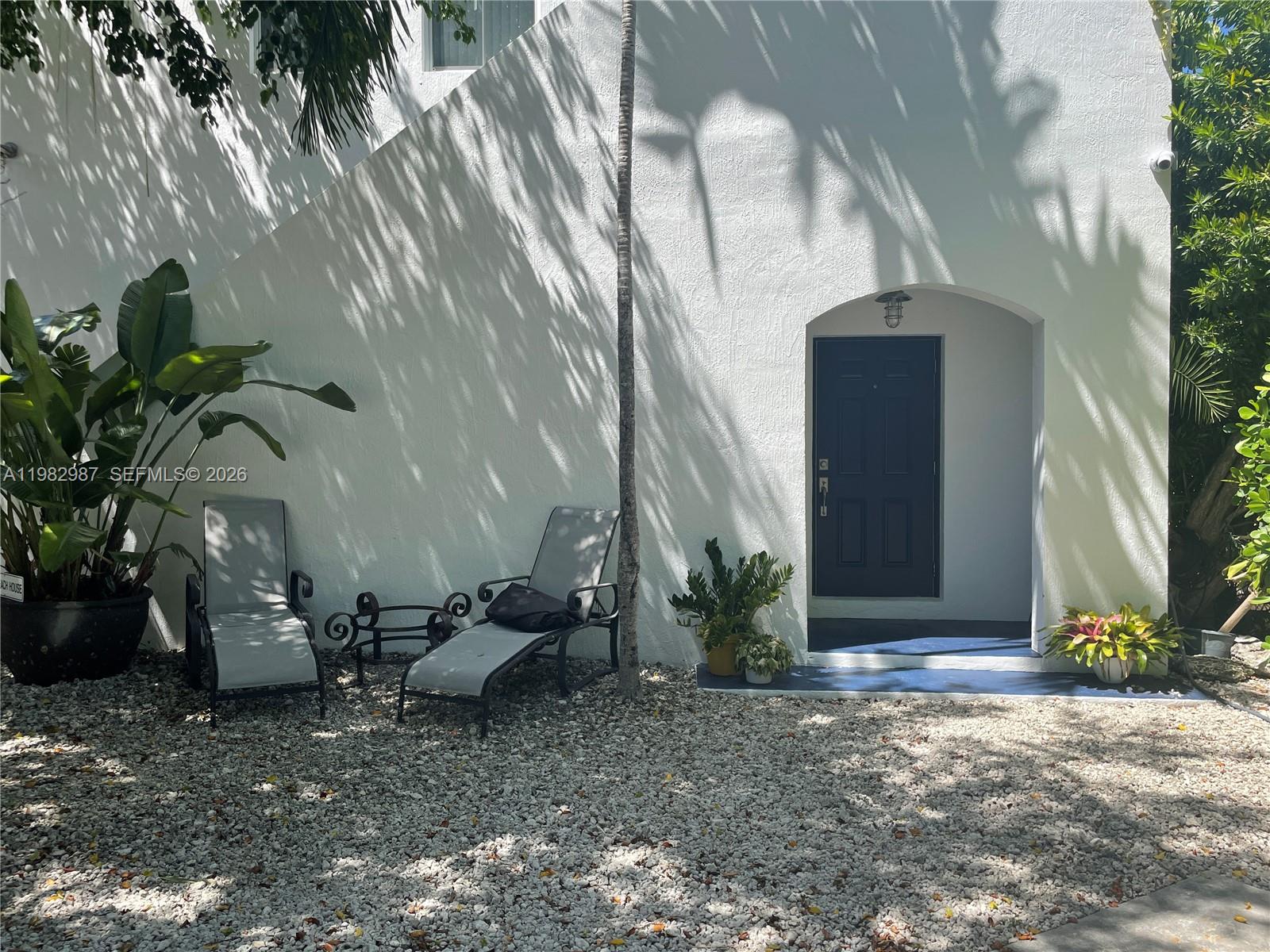 A white exterior wall features a dark door, two lounge chairs, and potted plants — 3421 SW 25th St # 01, Miami FL 33133, MIAMI, FL