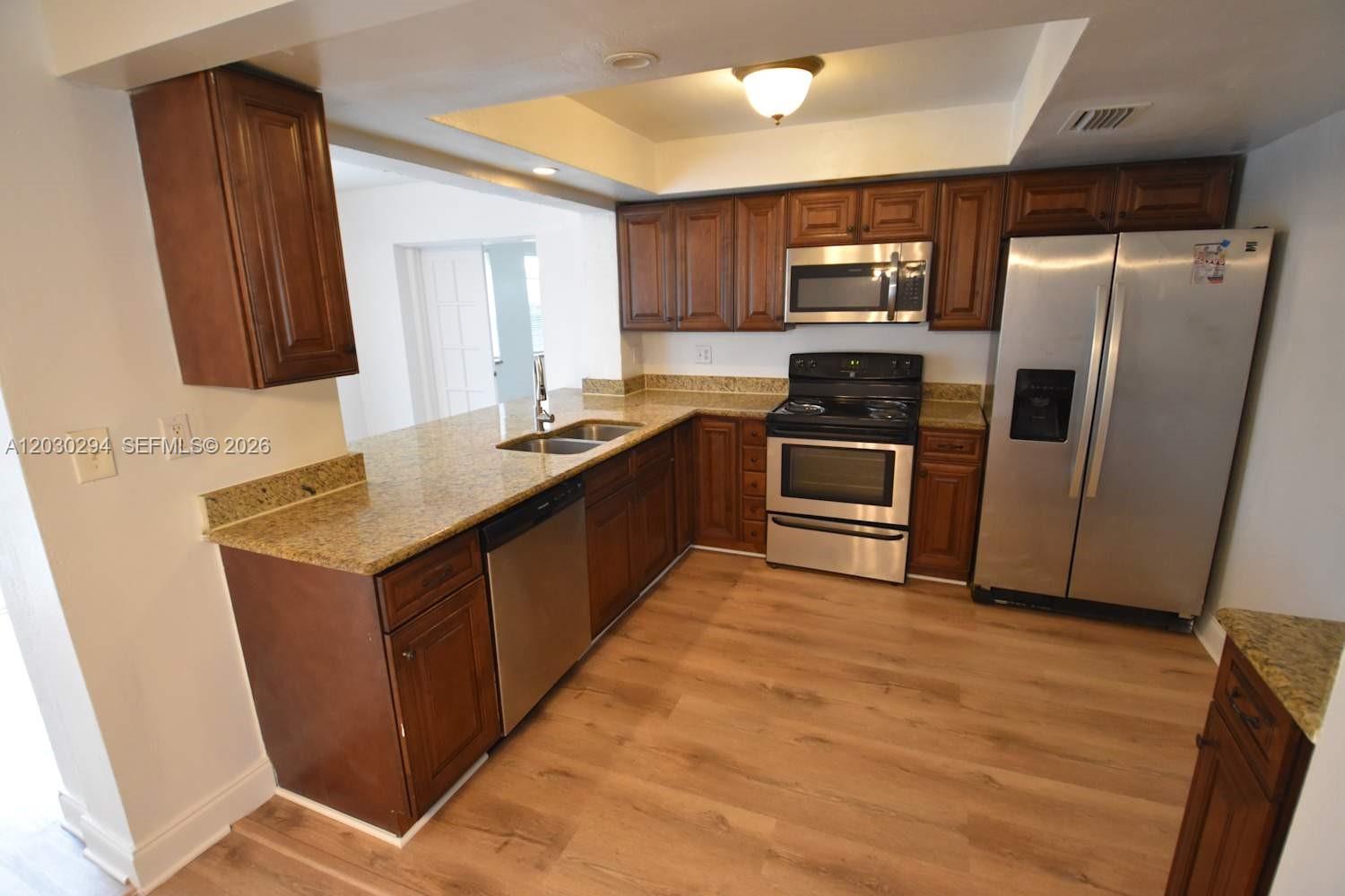A kitchen features wood cabinets, granite countertops, stainless steel appliances, and light wood-look flooring — SUNRISE, FL