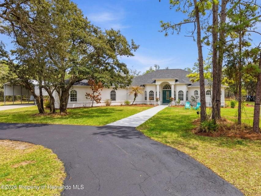 A white single-story house with a dark roof and a paved driveway sits among trees — 12124 SQUEEZE AVENUE, BROOKSVILLE, FL