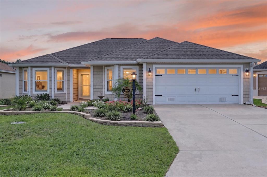 A single-story house with tan siding, a two-car garage, and a landscaped front yard at sunset — 1613 PENNECAMP DRIVE, THE VILLAGES, FL