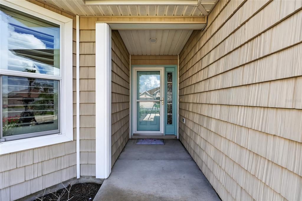 A covered entryway features tan shingle siding, a light blue front door, and a concrete porch — 1613 PENNECAMP DRIVE, THE VILLAGES, FL