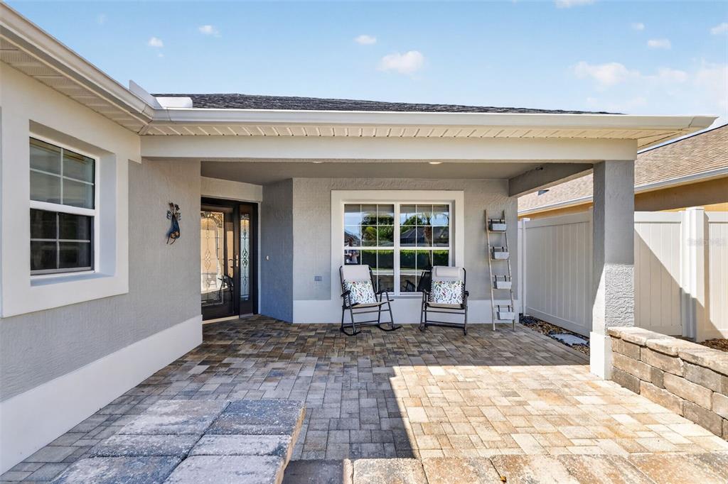 A covered patio features paver flooring, two rocking chairs, and a white vinyl privacy fence — 562 AVECILLA DRIVE, THE VILLAGES, FL