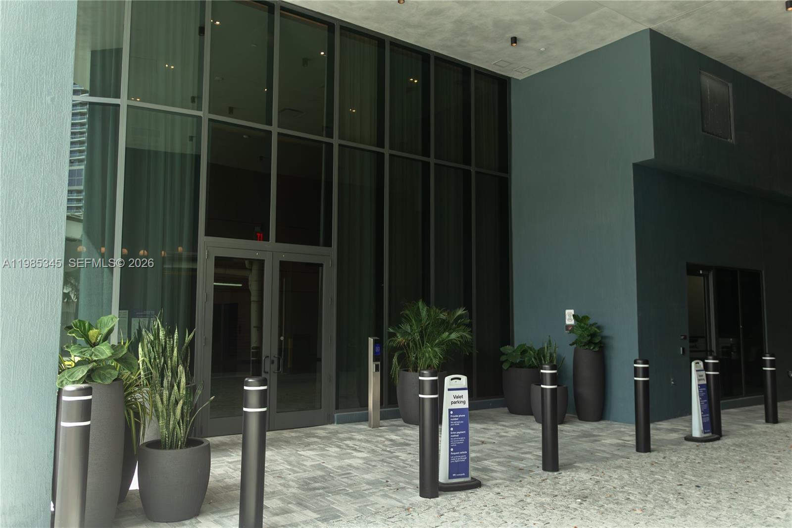 A modern building entrance features a glass facade, dark pillars, and potted plants on pavers — 121 NE 5th St # 2211, Miami FL 33132, MIAMI, FL
