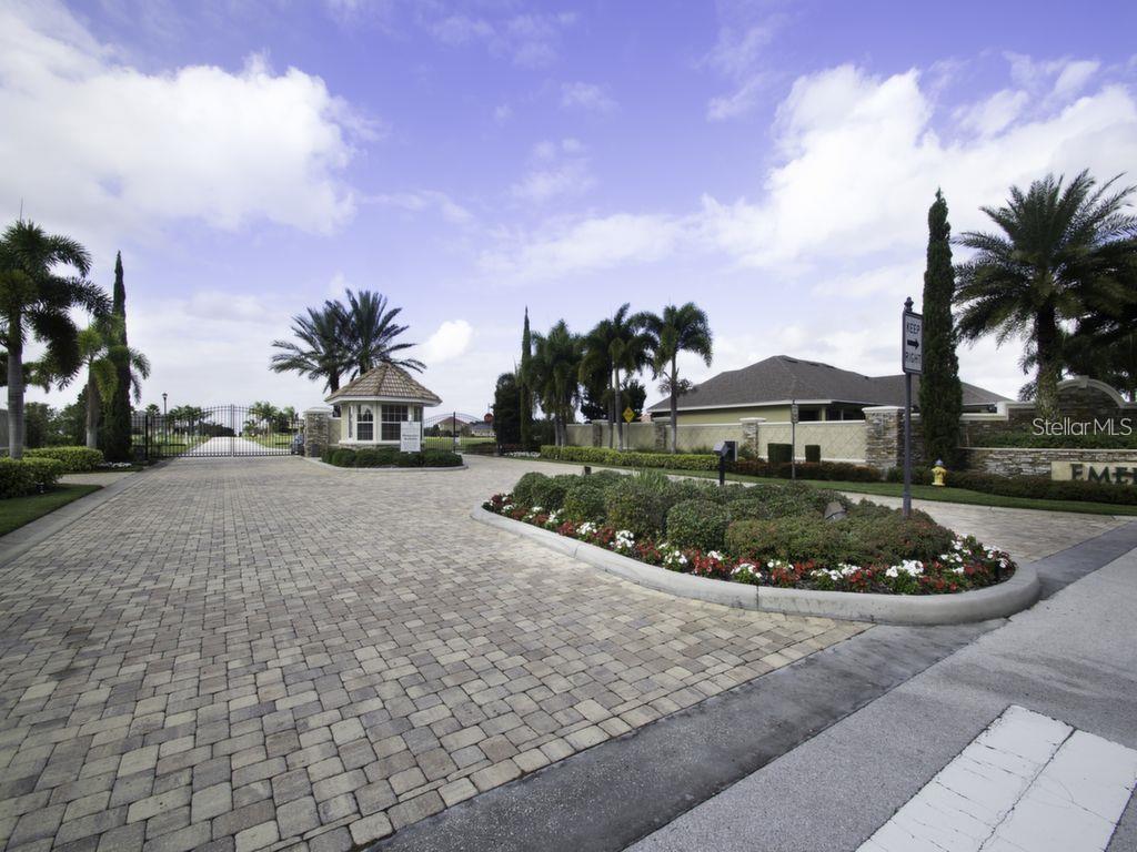 A gated entrance with a guardhouse, paved driveway, and landscaped median under a blue sky — 4410 EMERALD PALMS LANE, WINTER HAVEN, FL