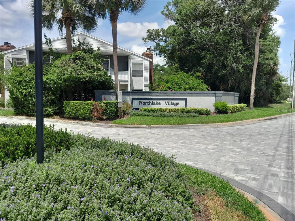 A stone monument sign for Northlake Village stands near a paved road and residential building — 25210 NORTHLAKE DRIVE UNIT 210, SANFORD, FL